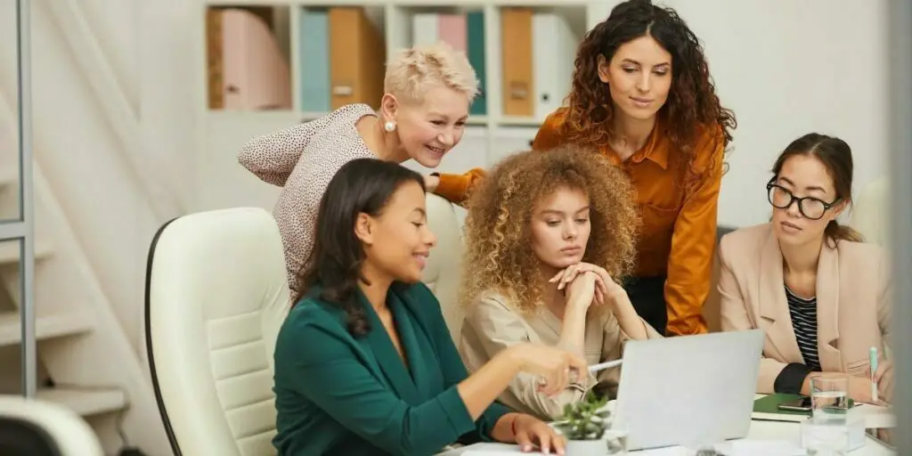 group of five positive stylish women using a laptop to work on projects during a business meeting