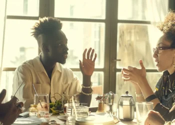 a diverse group of people sitting around a table, engaging in a thoughtful discussion with hands gestures and expressions of collaboration.